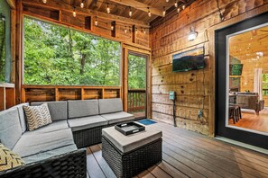 Interior de una cabaña de madera con sofá, televisor montado en la pared y un ventanal con vistas a la vegetación.