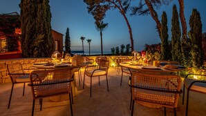 Un área de comedor al aire libre con sillas y mesas de mimbre, con vistas panorámicas al mar y árboles.