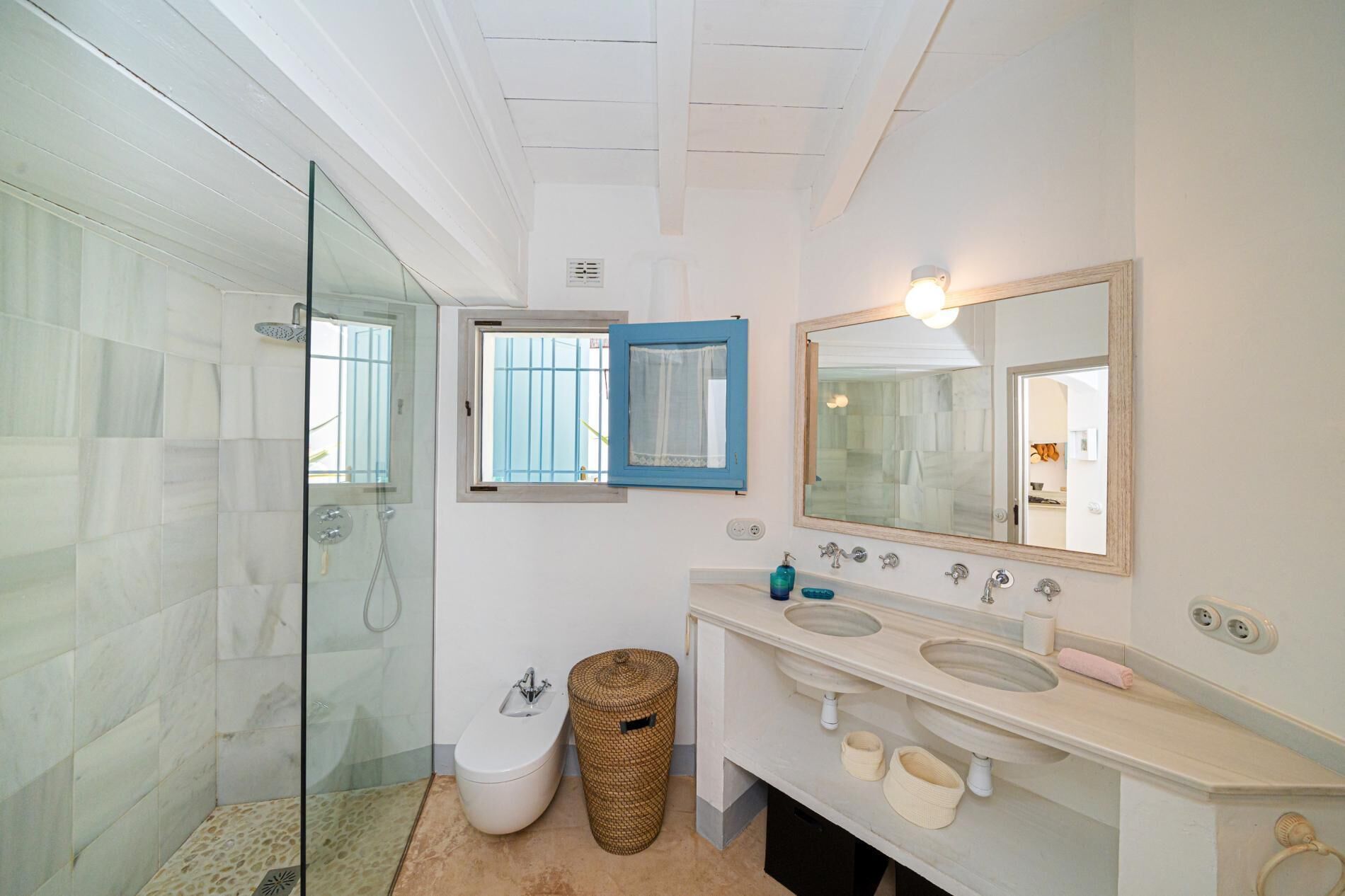 Une salle de bain avec une douche à parois de verre, des toilettes, un bidet, un lavabo avec un miroir et une fenêtre avec un cadre bleu.