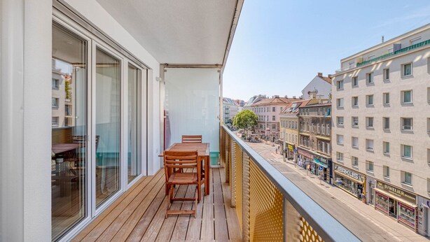 A balcony with a wooden table and chairs, a glass door, and a view of a city street with buildings and shops. - Sunny Brigittenau 1BR w/ Elevator & Storage, nr Augarten, by Blueground (Vienna)