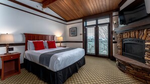A hotel room with a large bed, a stone fireplace, a television, and a window with blinds. - The Landing 211 Deluxe Queen Suite,  Jetted Tub (Chelan)