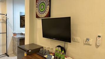 Quarto de hotel com televisão de tela plana na parede, cafeteira, e uma mesa pequena com controle remoto, garrafas de água e caixa de lenços.