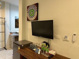 Quarto de hotel com televisão de tela plana na parede, cafeteira, e uma mesa pequena com controle remoto, garrafas de água e caixa de lenços.