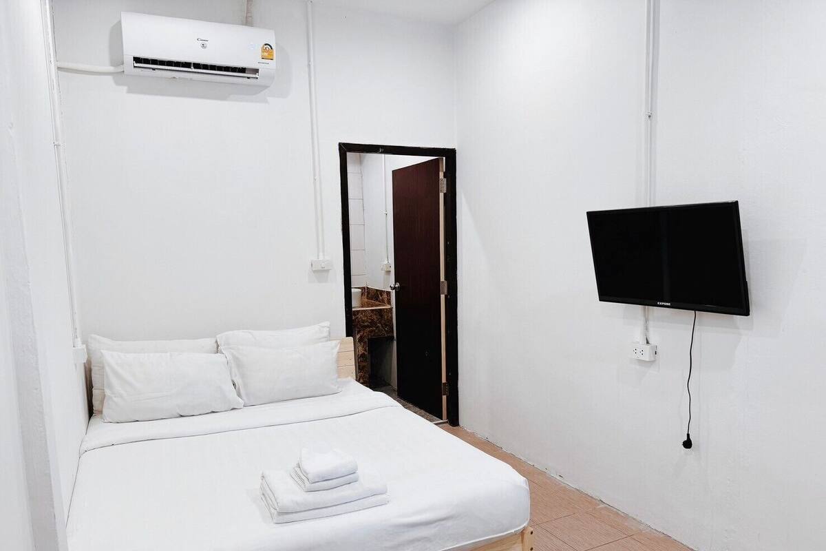 A clean, modern hotel room with a bed, white linens, a flat-screen TV mounted on the wall, and an air conditioning unit above the bed.