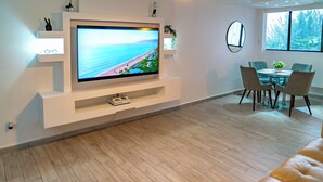 Un soggiorno moderno con televisore a schermo piatto, zona pranzo con tavolo e sedie in vetro, e un'ampia finestra con vista sull'esterno.