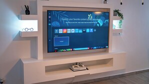 Un soggiorno moderno con una televisione a schermo piatto montata su una parete bianca, una console per videogiochi su un ripiano inferiore e oggetti decorativi sugli scaffali.