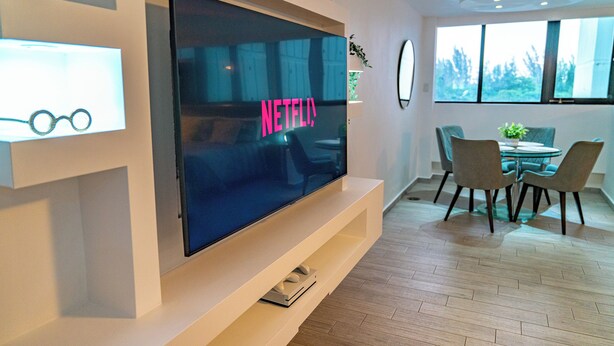 Un soggiorno moderno con un'ampia TV a schermo piatto montata su una parete bianca, un'area pranzo con un tavolo rotondo e sedie, e un pavimento in legno.