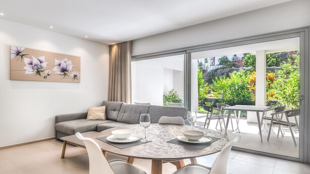 A modern living room with a grey sofa, a round dining table with white chairs, and a large window offering a view of a garden. - Colinas - Ancor 0.3 POOL VIEW 1B (Arona)