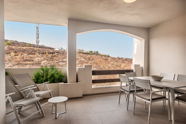 En moderne udendørs terrasse med spiseplads, inklusiv stole og et bord, med udsigt over et bakket landskab.