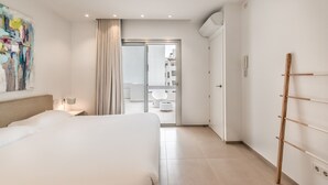 Une chambre d’hôtel moderne dotée d’un grand lit, d’une armoire, d’une salle de bain visible à travers une porte en verre et d’un balcon avec des meubles d’extérieur.