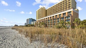 Ein Hotel direkt am Strand mit mehreren Etagen, ein Sandstrand im Vordergrund und ein klarer Himmel.