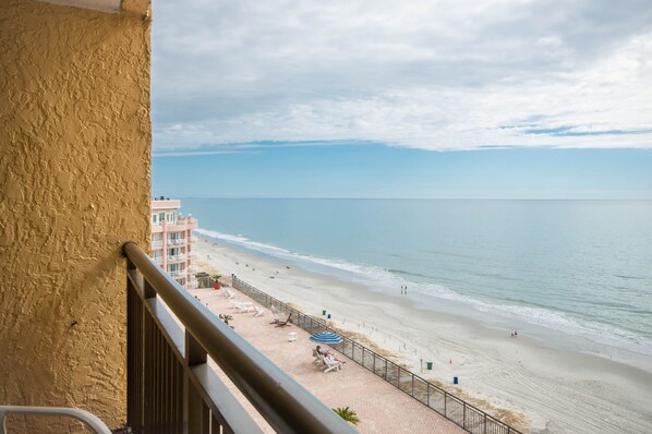 Vista al mar desde un balcón con barandilla, una playa arenosa y un edificio al fondo.