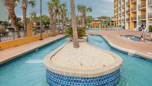 Ein Schwimmbecken mit Fliesenrand, Palmen und ein mehrstöckiges Hotel im Hintergrund.