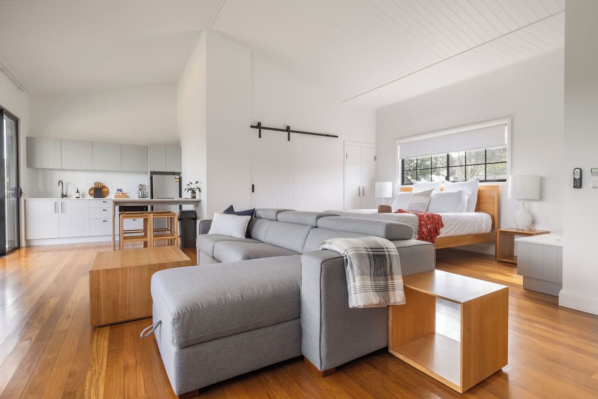 A modern living space with a grey sectional sofa, a wooden coffee table, a bed with white bedding, and a kitchen area with white cabinets.