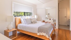 Ein modernes Schlafzimmer mit einem hölzernen Bett, weißer Bettwäsche, einer Nachttischlampe und Blick ins Grüne durch ein Fenster.