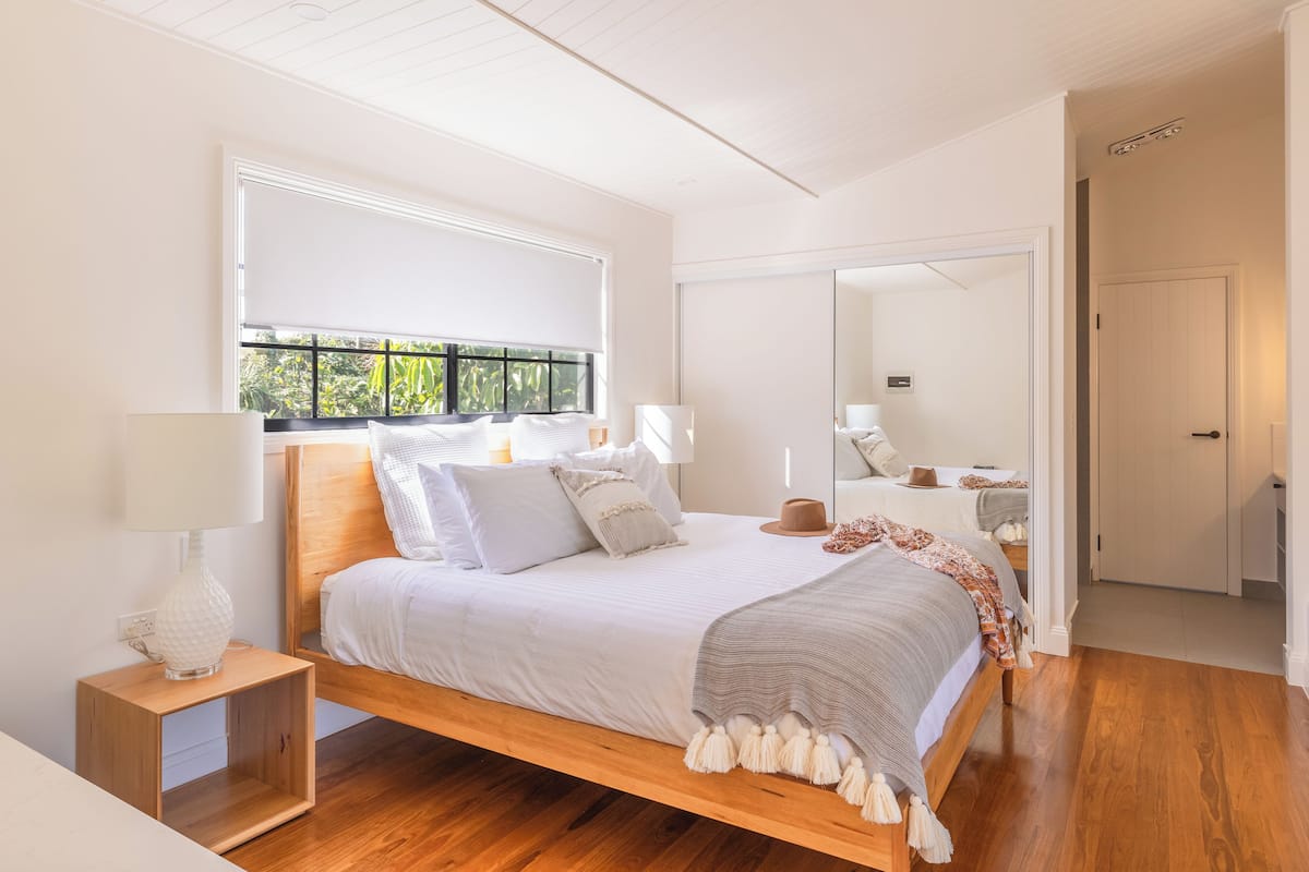 A modern bedroom with a wooden bed, white bedding, a bedside lamp, and a view of greenery through a window.
