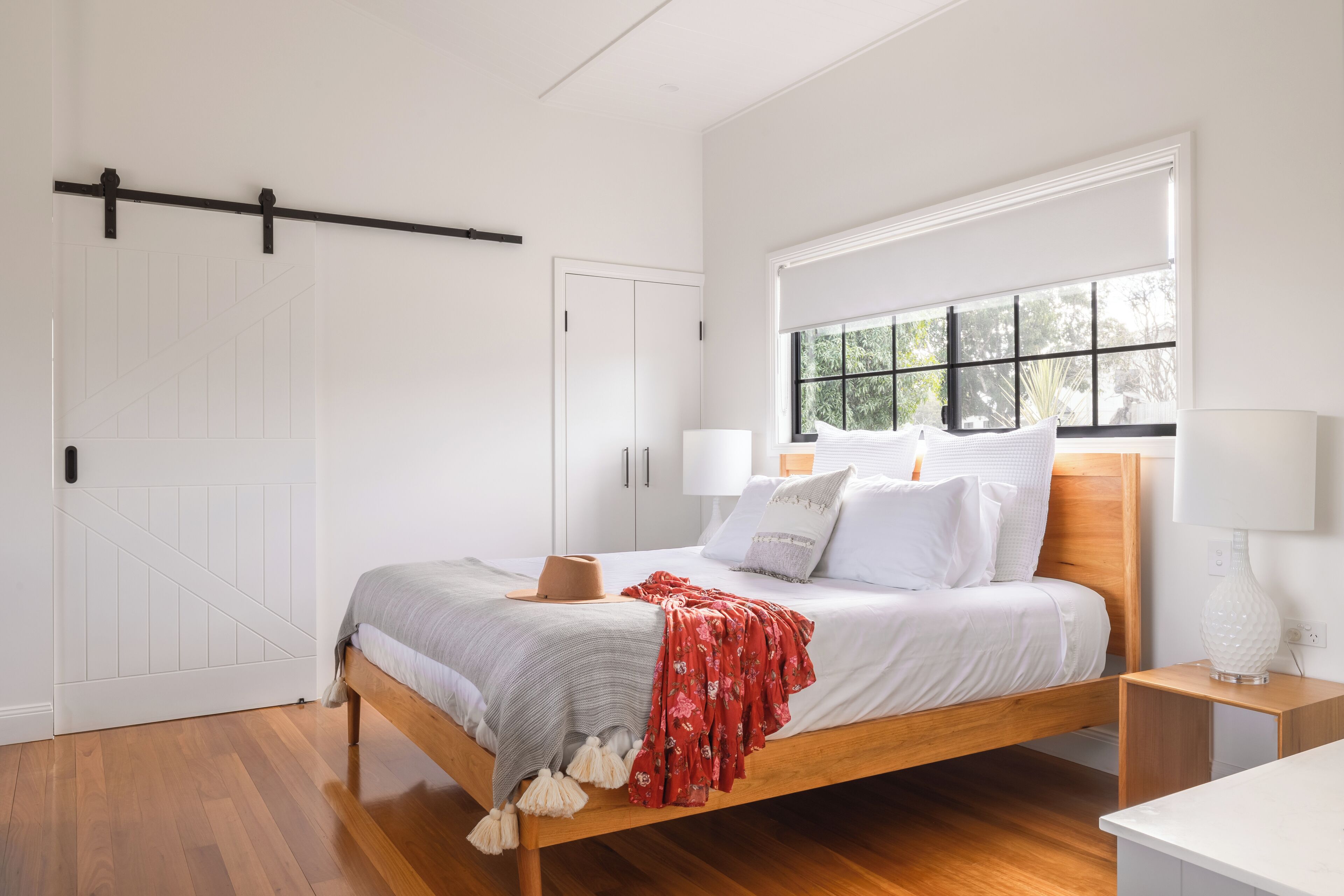 Un dormitorio con una cama de madera, paredes blancas, una ventana con persianas, una mesita de noche con una lámpara y una puerta con sistema corredizo.