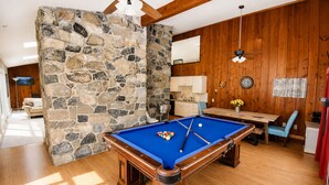 A pool table in a room with wooden flooring, stone walls, a wooden ceiling, and a dining area with a table and chairs. - Large North Conway Vacation Home Indoor Pool, Pool Table Sleeps 22 (Conway)