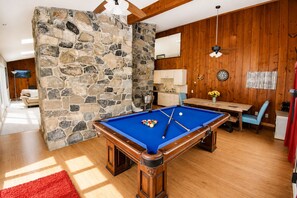Una mesa de billar en una sala con piso de madera, paredes de piedra y techo de madera, además de un comedor con una mesa y sillas.