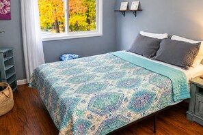 Une chambre à coucher avec un lit, des oreillers, une fenêtre donnant sur des arbres aux couleurs automnales, une étagère avec des photos et un plancher en bois.