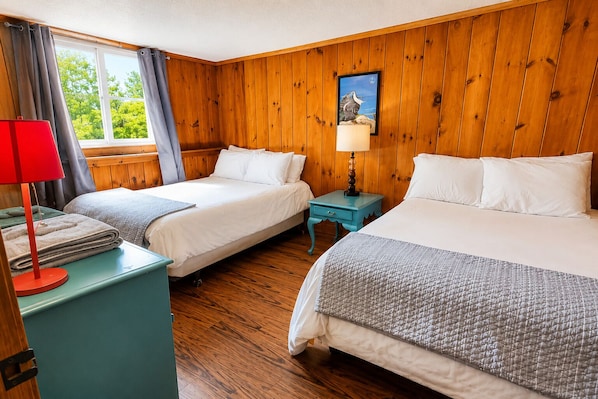 A room with two beds, a window with curtains, a wooden wall, and a small table with a lamp.