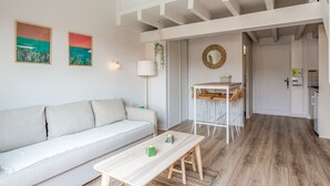 A modern living room with a white sofa, a wooden coffee table, and a dining area with a white table and chairs. There is a kitchen visible in the background.