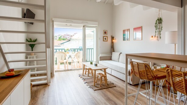 A modern living room with a wooden floor, white furniture, a balcony with outdoor seating, and a view of buildings in the distance.