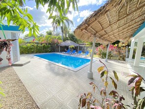 A swimming pool with lounge chairs, surrounded by tropical plants and a thatched roof structure.