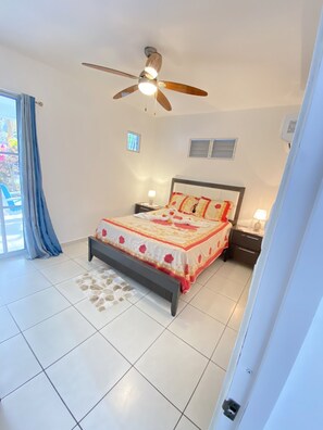 A bedroom with a bed, two bedside tables, a ceiling fan, a window, and a view of the outdoors.