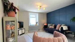 A modern living room with a curved sofa, a glass cabinet with decorative items, a blue accent wall, and a window providing natural light. - Harbourside - Luxury 2Bed Cottage & Garden (Weymouth)