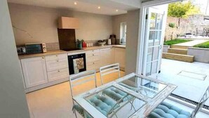 A modern kitchen with white cabinets, a built-in oven, and a dining area with a glass table and blue cushioned chairs. - Harbourside - Luxury 2Bed Cottage & Garden (Weymouth)