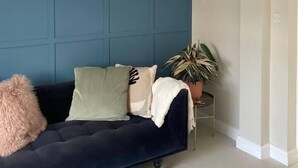 A room with a dark blue accent wall, a dark blue sofa with multiple cushions, a white ceiling with a gold chandelier, and a potted plant. - Harbourside - Luxury 2Bed Cottage & Garden (Weymouth)