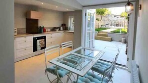 A modern kitchen with white cabinets and a glass dining table with blue upholstered chairs. - Harbourside - Luxury 2Bed Cottage & Garden (Weymouth)