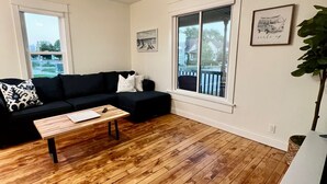 Sala de estar com sofá preto, piso de madeira, ventilador de teto e uma grande janela com vista para casas.