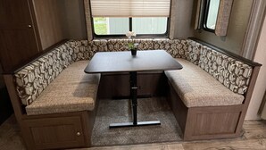 A compact RV interior with a dining table and bench seating, a window with blinds, and a mirror on the wall. - Spacious RV w/ large shower & Queen bed (Pacific)