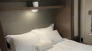 A neatly made bed with white linens, a small potted plant on a shelf above, and a wooden wardrobe to the right. - Spacious RV w/ large shower & Queen bed (Pacific)