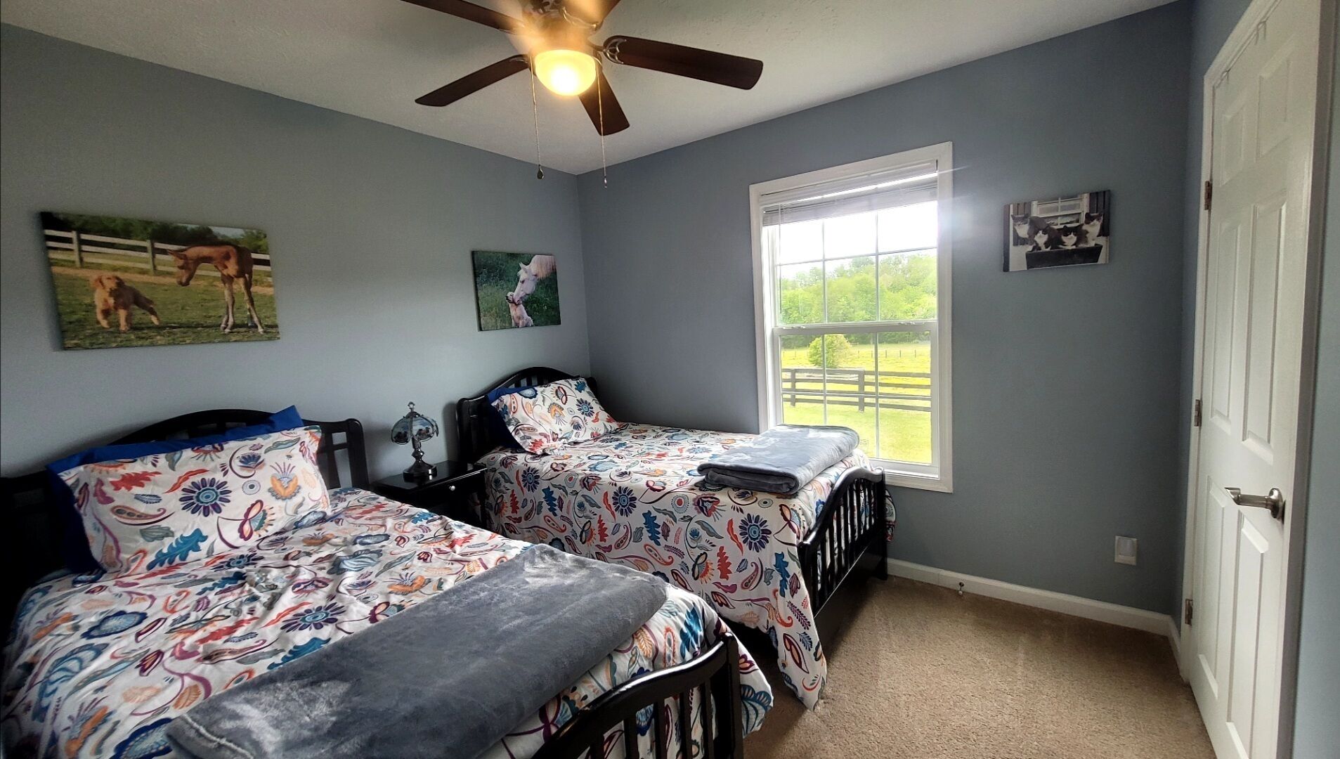 A bedroom with two beds, a ceiling fan, a window with a view of a fence, and a door.
