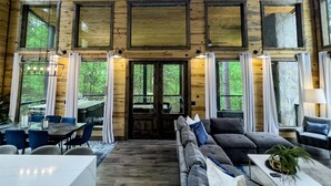 A spacious wooden interior with a dining area, seating, and large windows offering views of the forest.