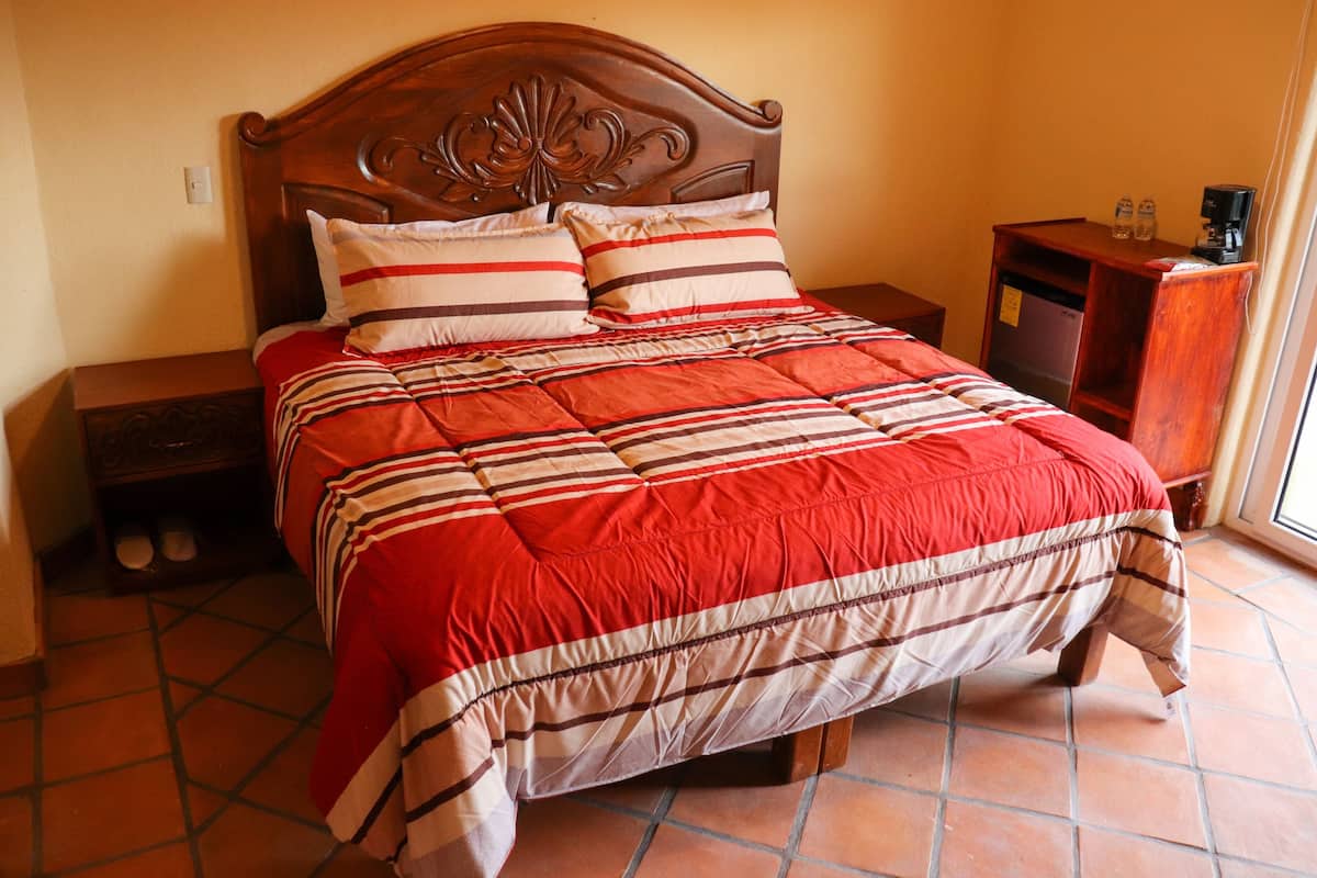 A neatly made bed with a striped bedspread and two pillows, a wooden headboard, a nightstand with a lamp, and a small wooden cabinet with a coffee maker.