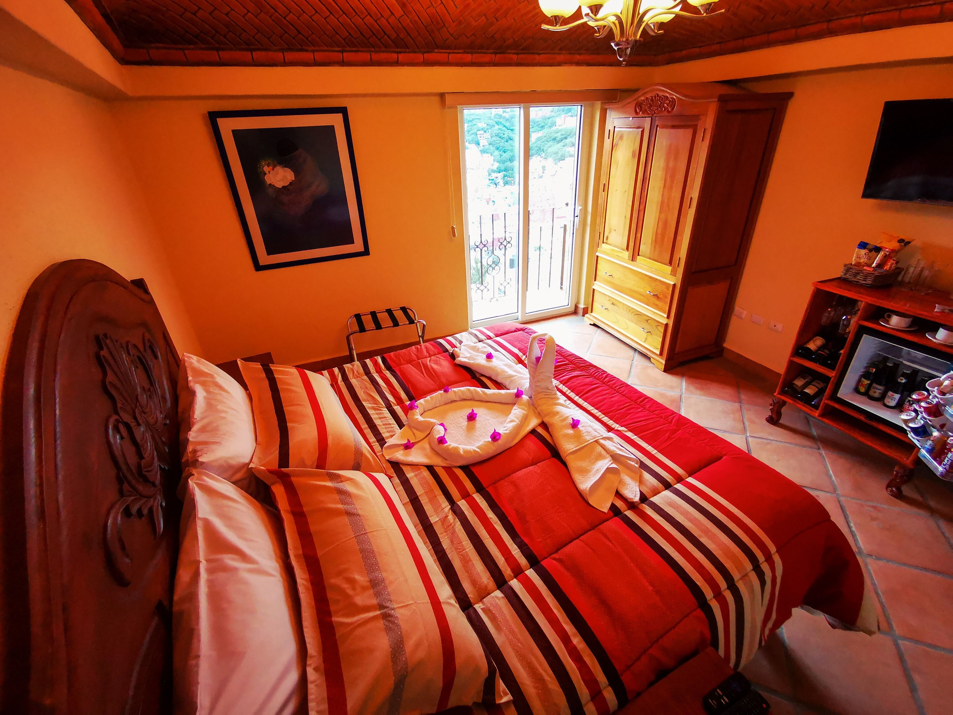 Een hotelkamer met een bed, een houten hoofdbord, een schilderij aan de muur, een houten kastje met flessen, en een raam met uitzicht op groen.