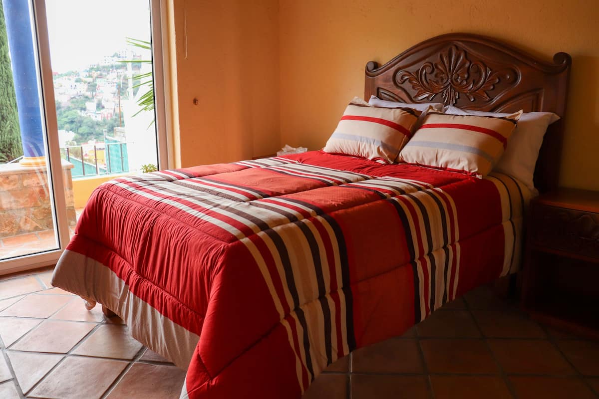 A bed with a striped blanket, pillows, and a wooden headboard in a room with a tiled floor and a view through a window.