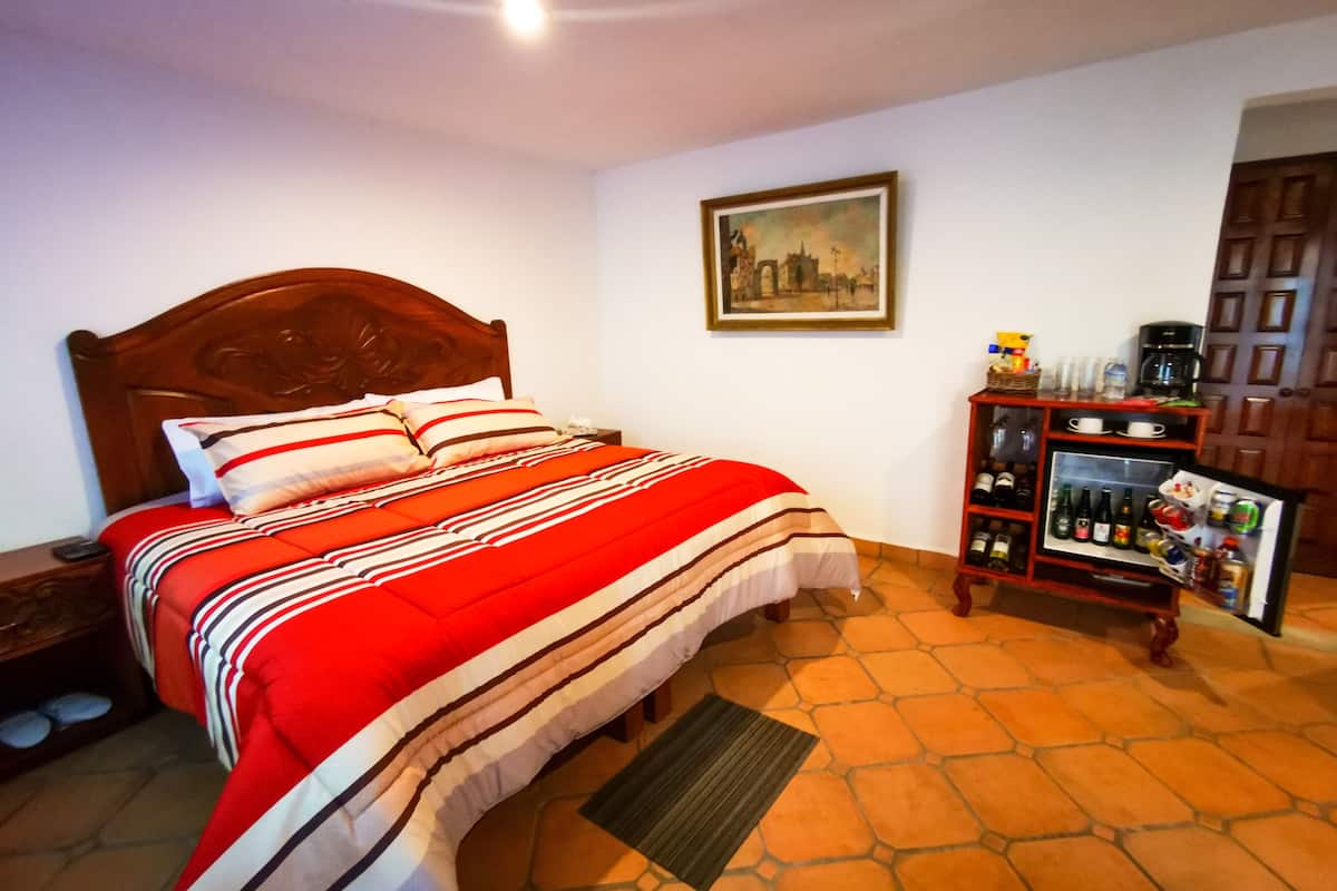 A neatly made bed with a striped blanket, a wooden headboard, a nightstand with a phone, a framed picture on the wall, and a minibar with various items.