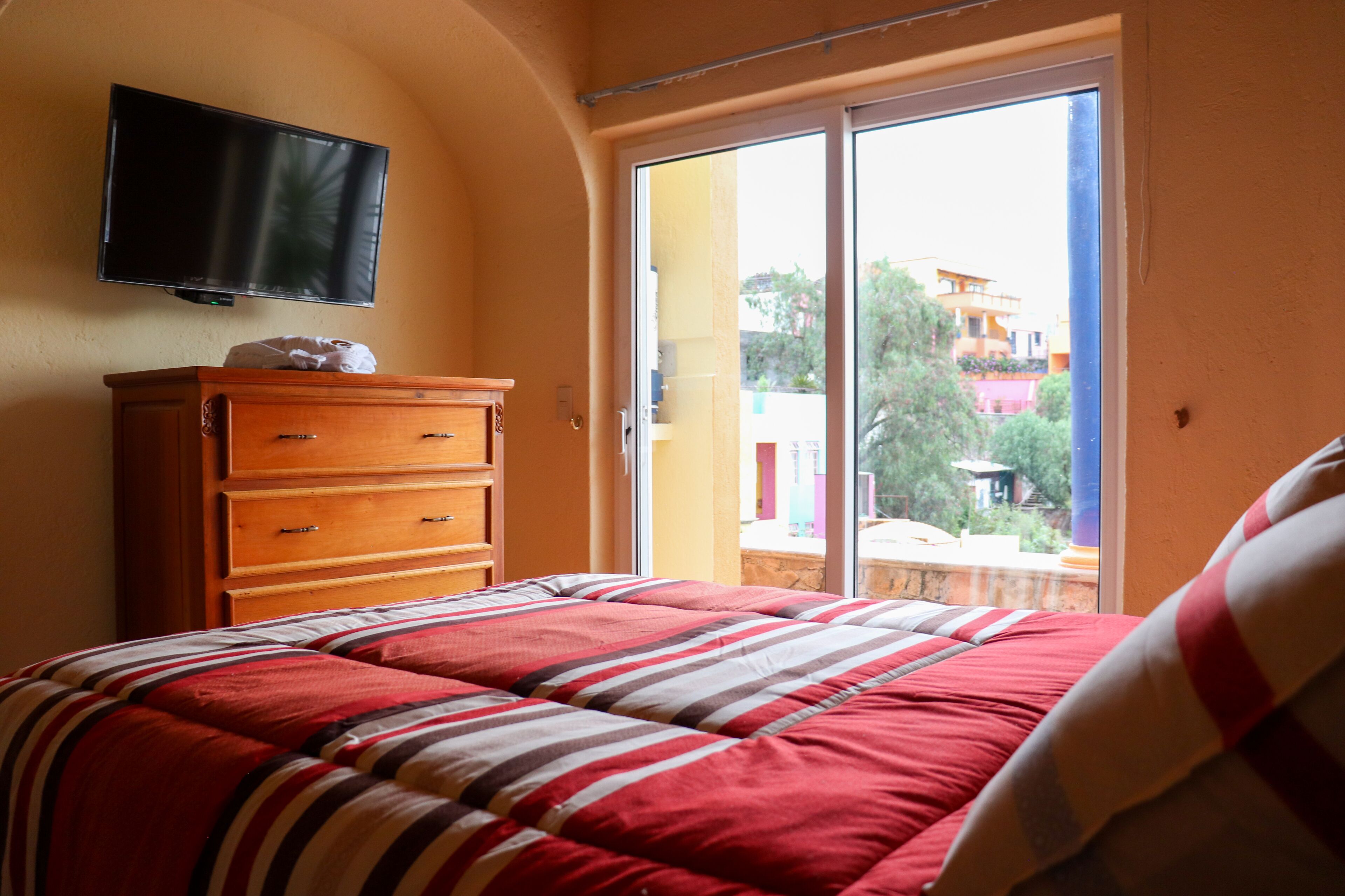 Ein Schlafzimmer mit Bett, Kommode, Fernseher an der Wand und einer Schiebetür zu einem Balkon.