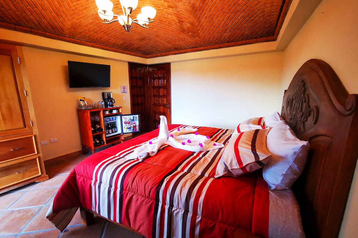 A bedroom with a brick ceiling, a bed with striped bedding, a television on the wall, and a small refrigerator.