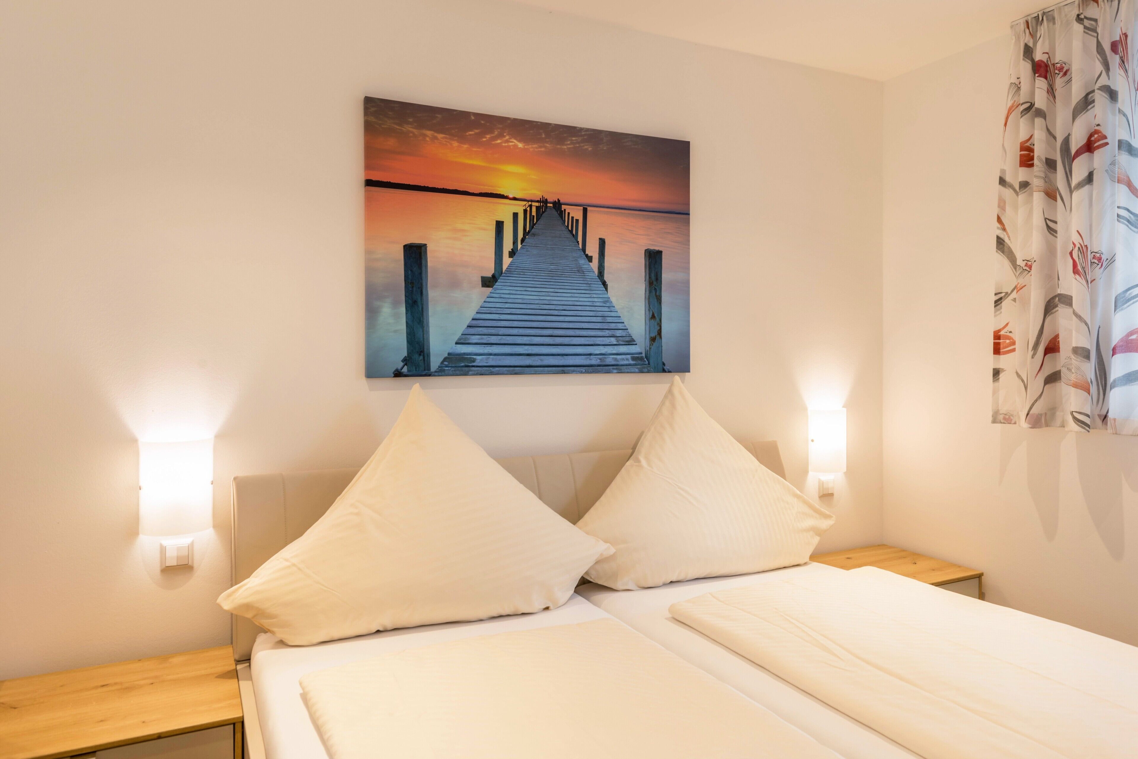Ein Hotelzimmer mit einem Bett, zwei Kissen, einem Nachttisch, einem Wandbild von einem Pier und einem Vorhang mit einem floralen Muster.