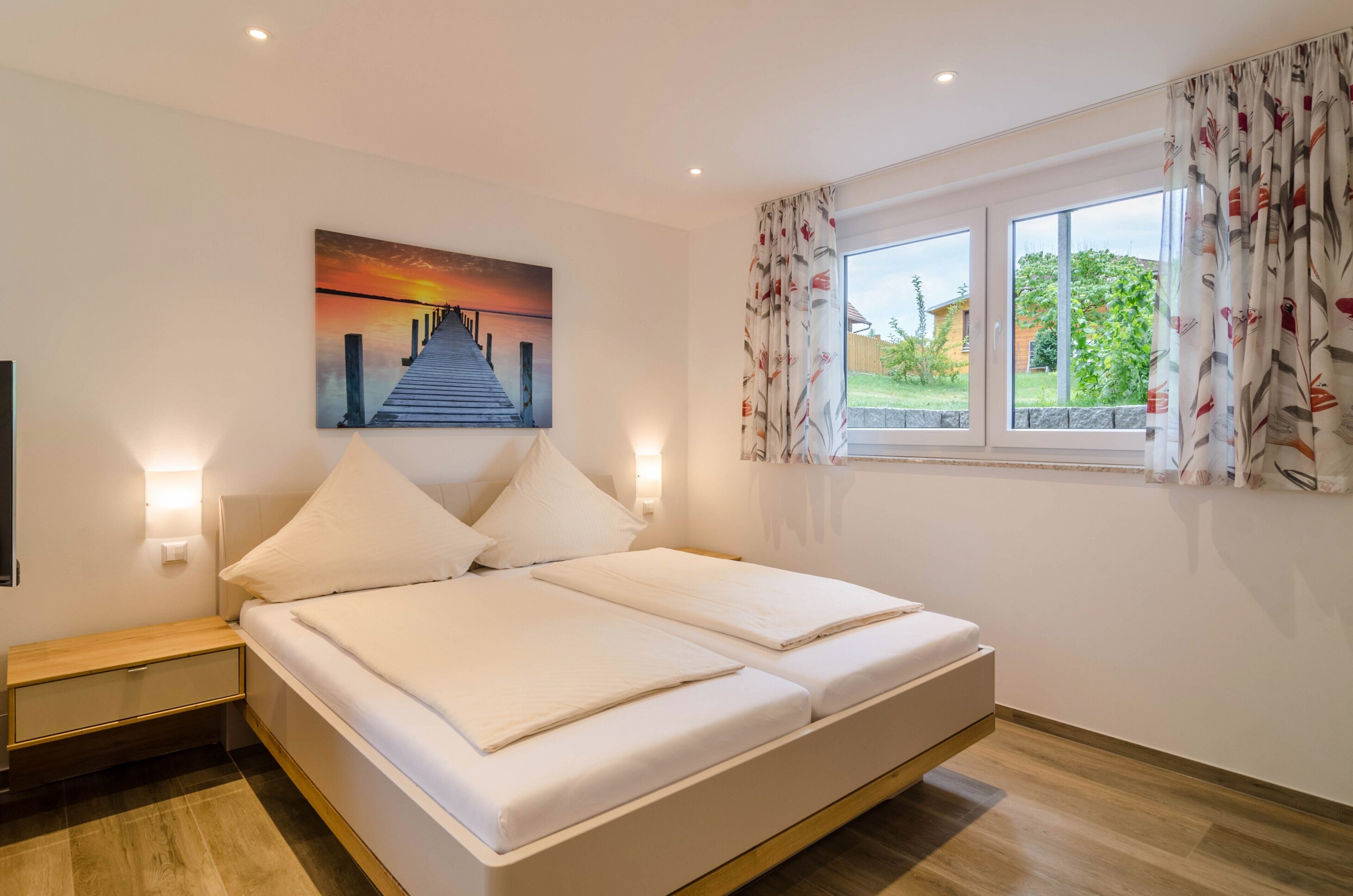 A double bed with a headboard, a nightstand with a drawer, a painting of a sunset over a pier, and a window with curtains.