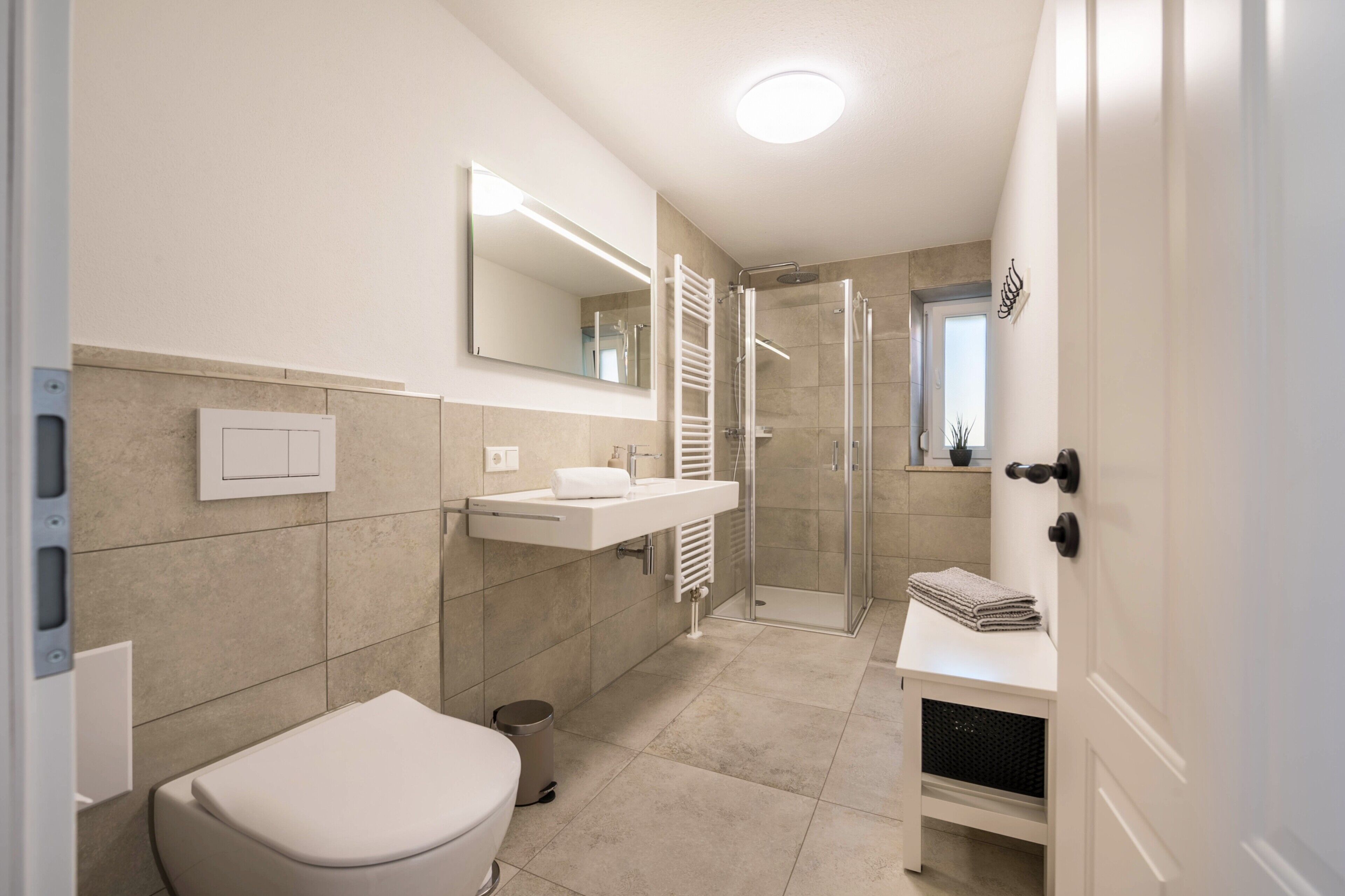 A modern bathroom with a toilet, sink, mirror, shower, and a small shelf with towels.