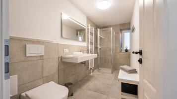 A modern bathroom with a toilet, sink, mirror, shower, and a small shelf with towels.