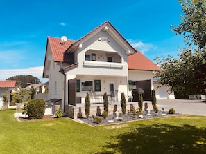 A two-story house with a red-tiled roof, a balcony, and a well-maintained lawn with small trees.