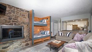 Un salon chaleureux avec une cheminée en pierre, un lit en mezzanine en bois, un canapé et une cuisine en arrière-plan.